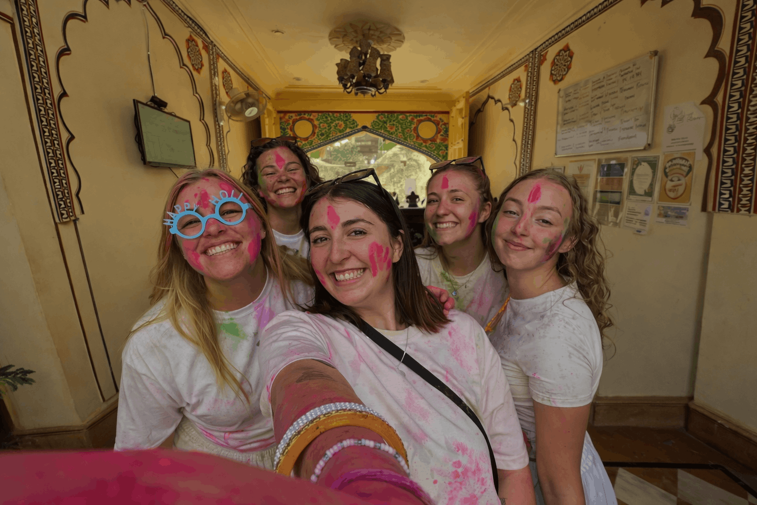 Tourists celebrating Holi in Rajasthan, smiling for a group selfie with faces covered in vibrant pink and green gulal inside a traditional heritage building